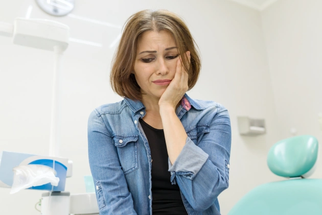 Woman in dental office examining teeth with hand to face, dental equipment visible in background.