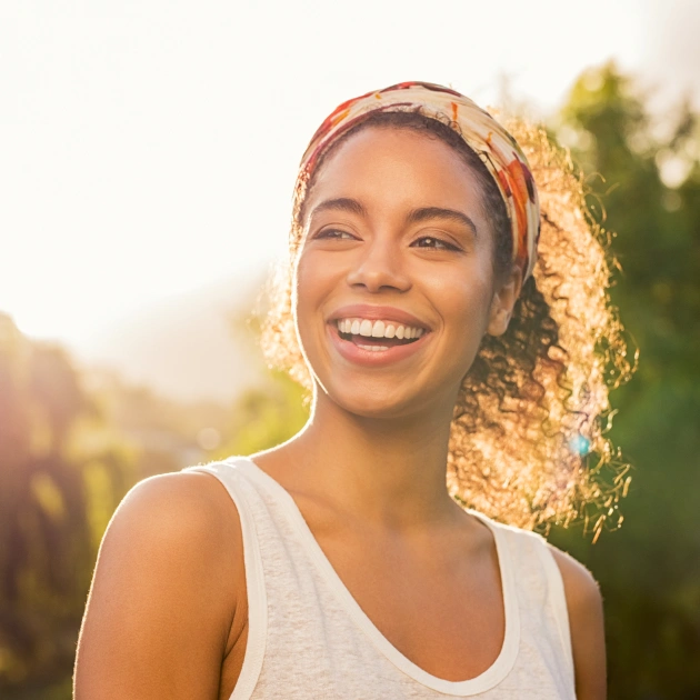 Smiling woman wearing white tank top and headscarf outdoors in sunny garden setting.