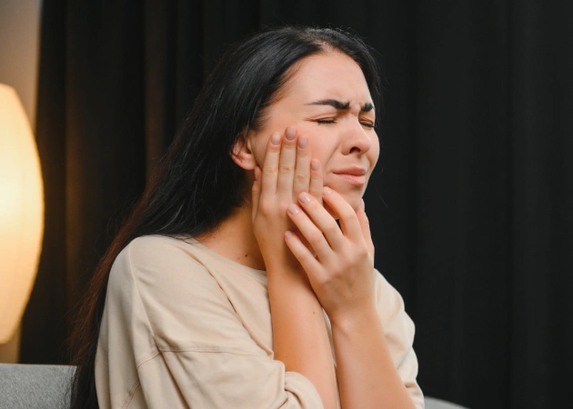 Woman touching jaw in pain, wearing beige shirt, indoor setting with warm lighting.