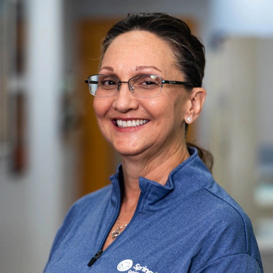 Smiling dental professional wearing glasses and blue scrubs in a clinical office setting.