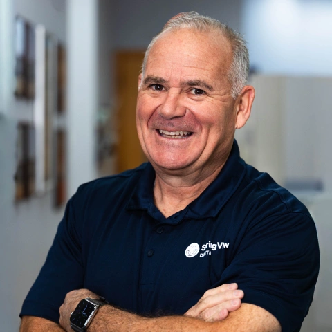 Smiling man in dark polo shirt with dental practice logo in professional office hallway setting.