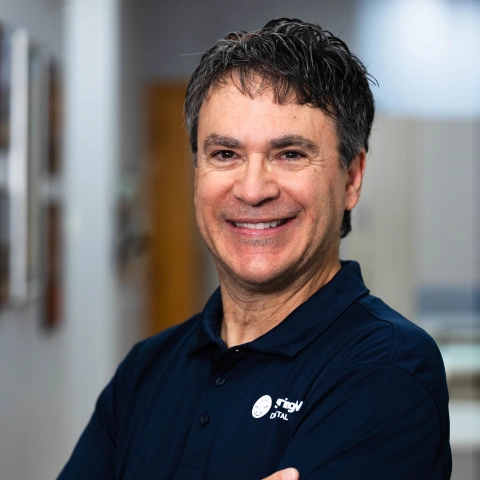 Professional headshot of smiling man in navy dental practice polo shirt, indoor office setting.